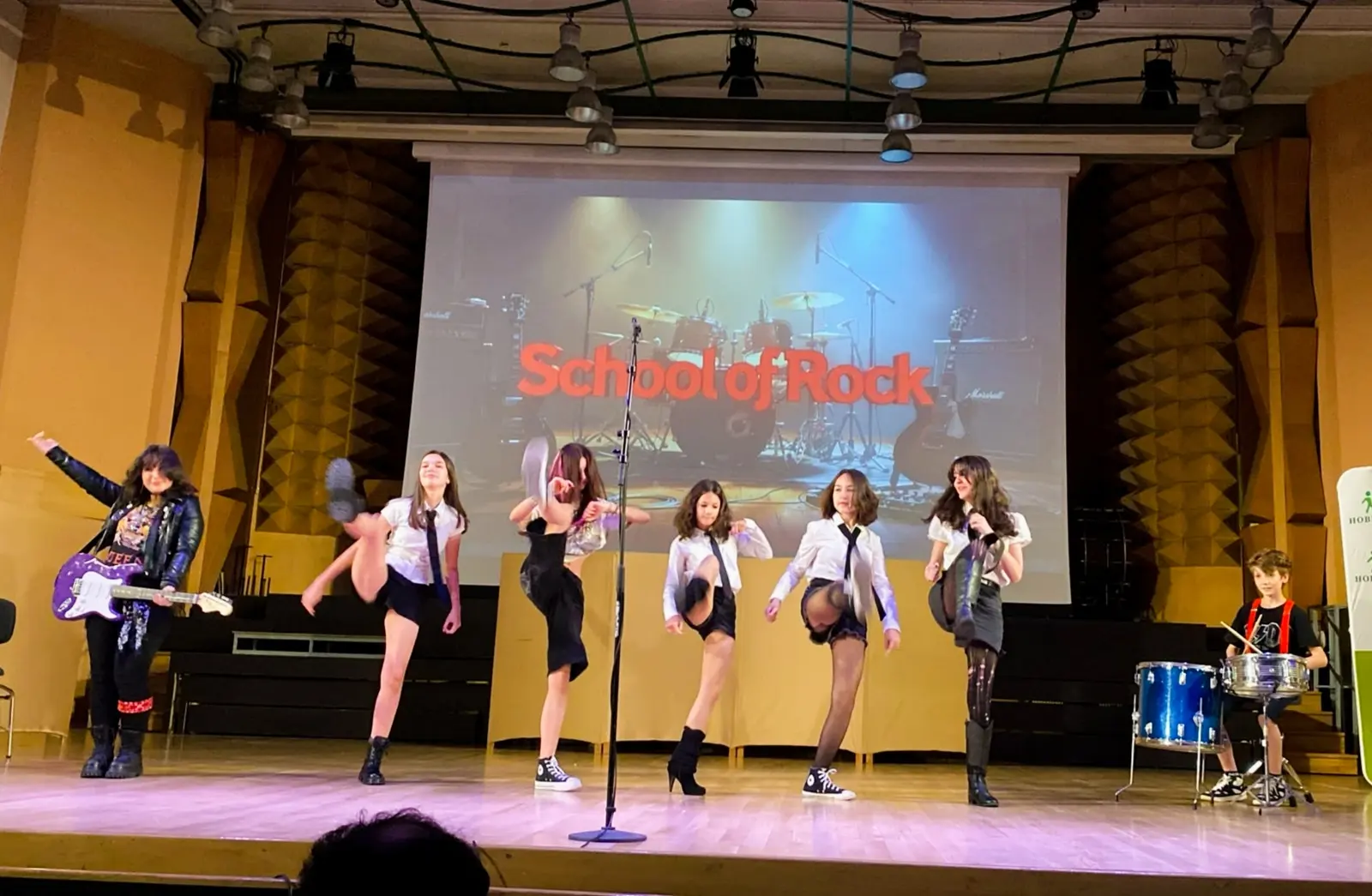 Elevii academiei pe scenă, cu microfoane și baterie, în spectacolul „School of Rock”