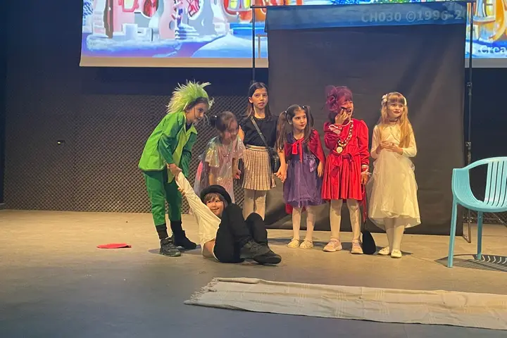 Grup de elevi în costume colorate, pe scenă, în fața unui decor de oraș de poveste