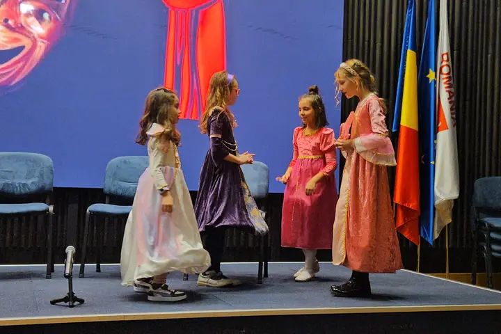 Eleve în rochii lungi, pe scena unei săli oficiale, lângă drapelele României și ale Uniunii Europene