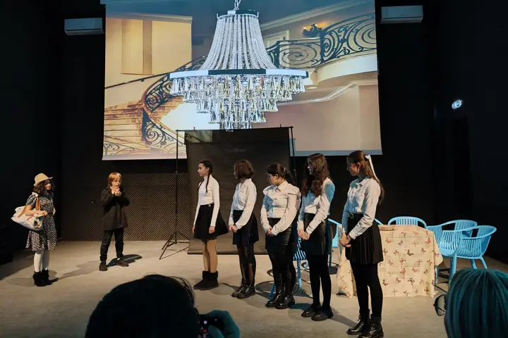 Grupă de eleve aliniate pe scenă, sub o proiecție cu un candelabru de palat
