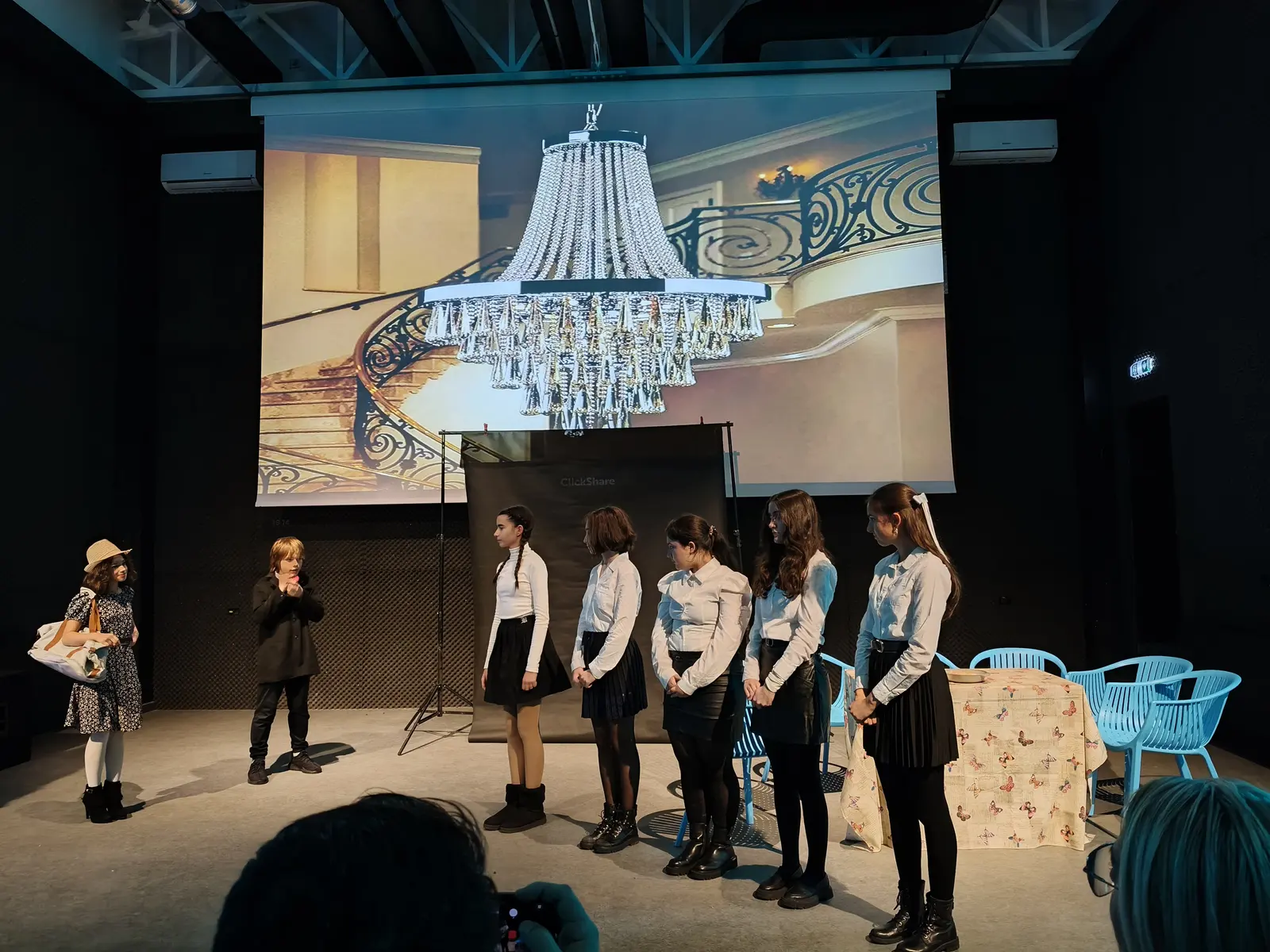 Grupă de eleve aliniate pe scenă, sub o proiecție cu un candelabru de palat