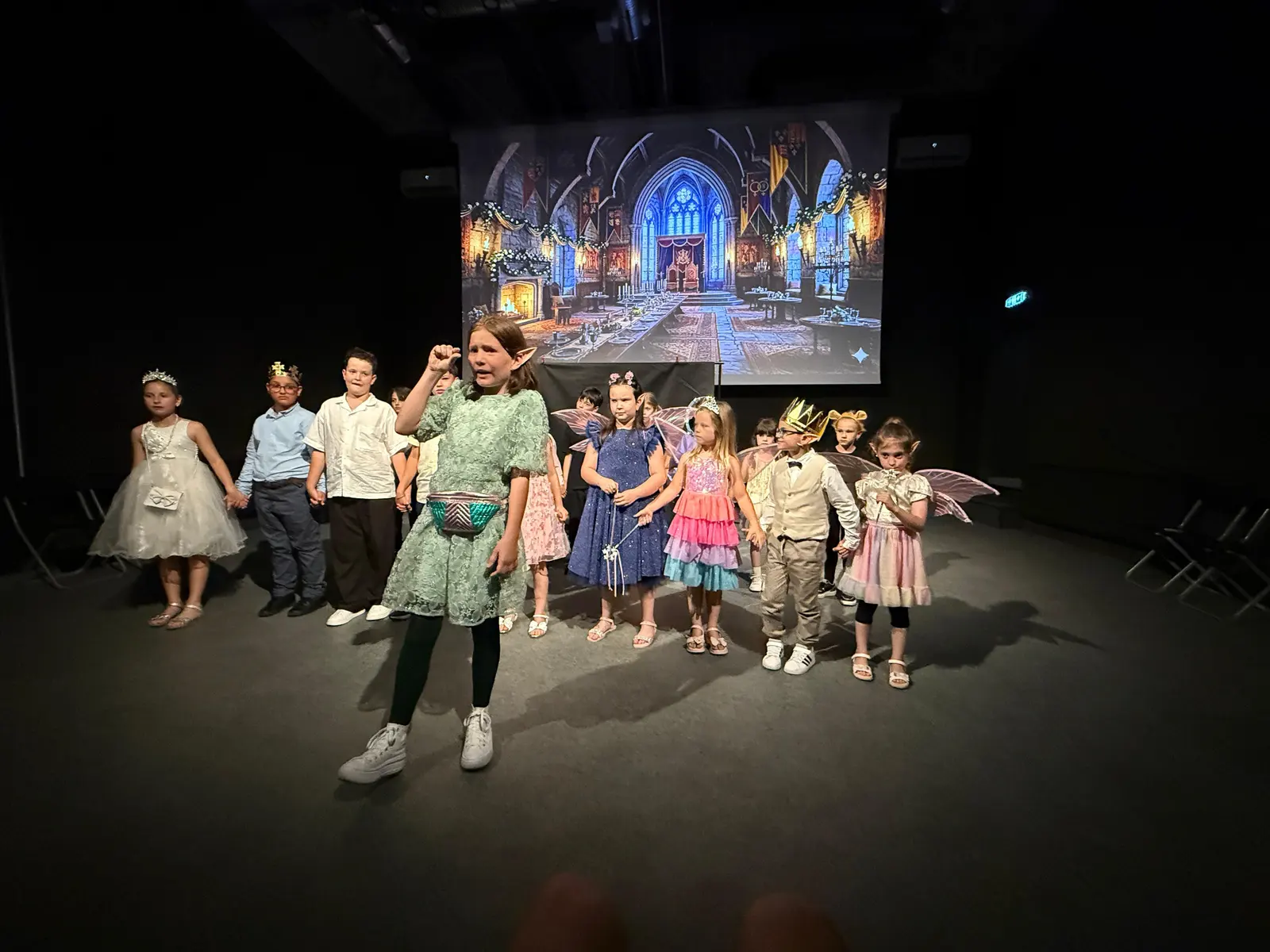 Grup de copii mici în costume de zâne, pe scenă, alături de profesoară